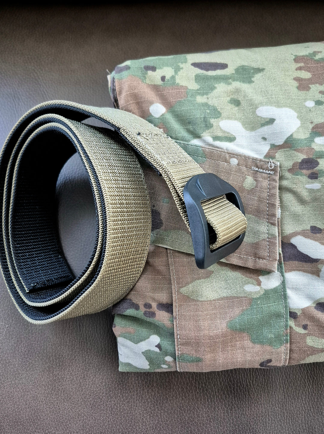 GS-004 Reversible Riggers Belt shown rolled and placed on camouflage-patterned fabric over brown surface; features matte black buckle and rugged nylon webbing, styled for tactical integration in field gear setups or outdoor deployment visuals