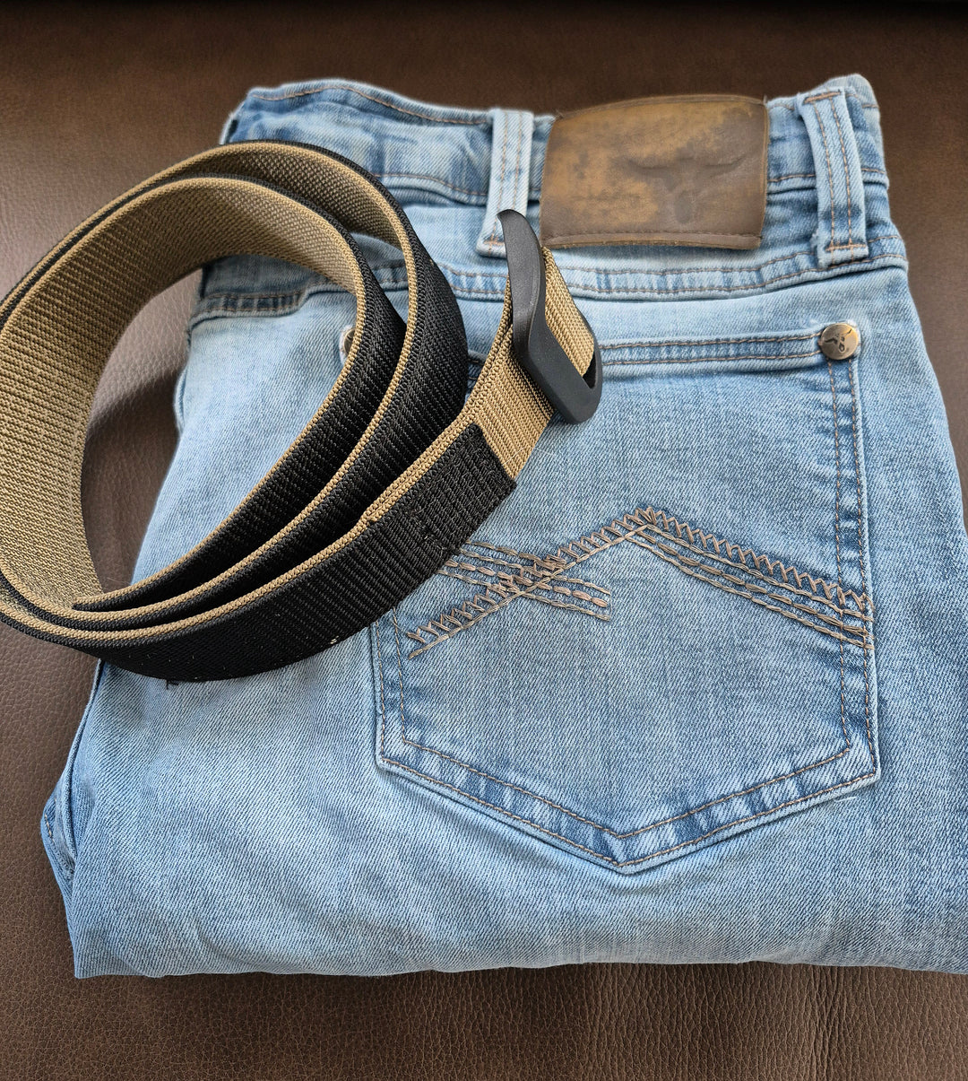GS-004 Reversible Riggers Belt coiled atop folded blue jeans on brown surface; belt features matte black buckle and black-coyote colorway, styled with visible rear pocket stitching and leather patch to suggest off-duty utility or rugged lifestyle pairing