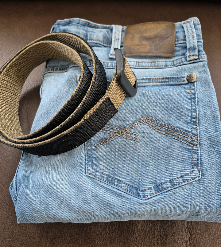GS-004 Reversible Riggers Belt coiled atop folded blue jeans on brown surface; belt features matte black buckle and black-coyote colorway, styled with visible rear pocket stitching and leather patch to suggest off-duty utility or rugged lifestyle pairing