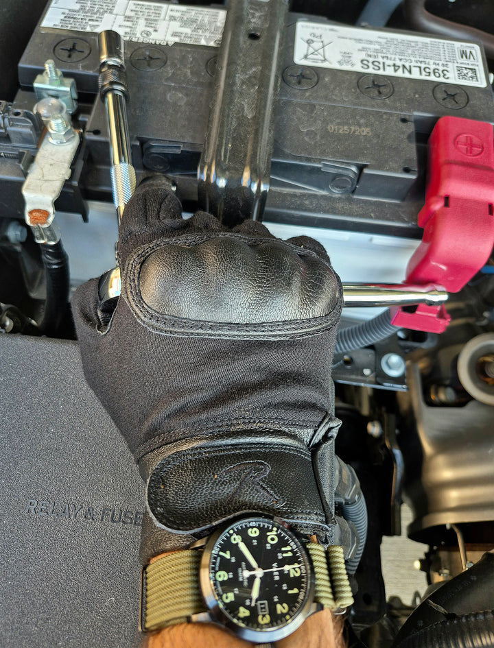 GS-005 Kevlar Tactical Gloves in use during automotive maintenance, shown gripping a wrench to adjust a battery terminal; black glove features molded knuckle protection and goatskin detailing, paired with wristwatch for rugged field utility
