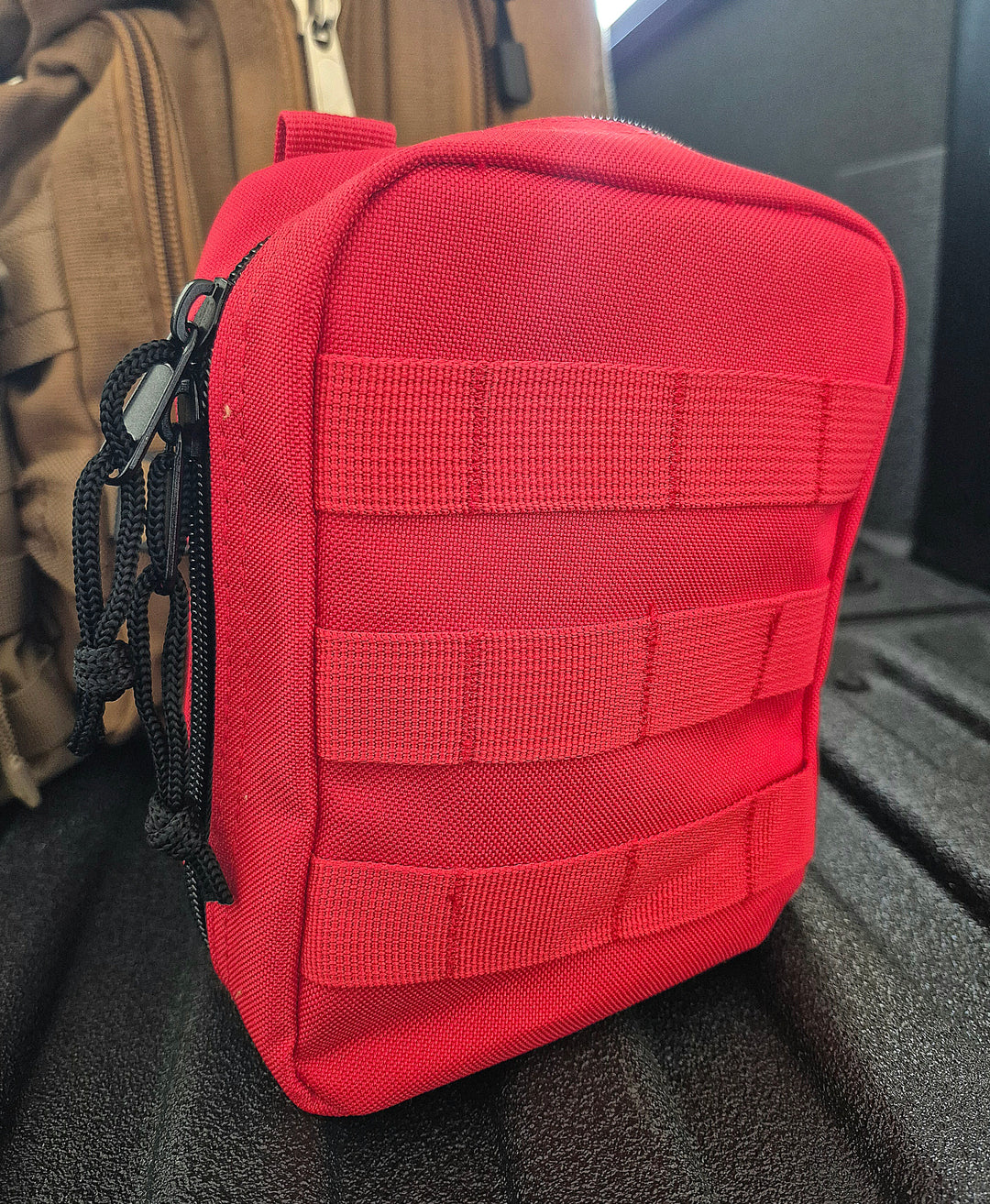 GS-010 Red Tactical Pouch displayed on black textured surface with horizontal webbing and black zipper cords; background features a brown carry bag, reinforcing rugged field gear aesthetic