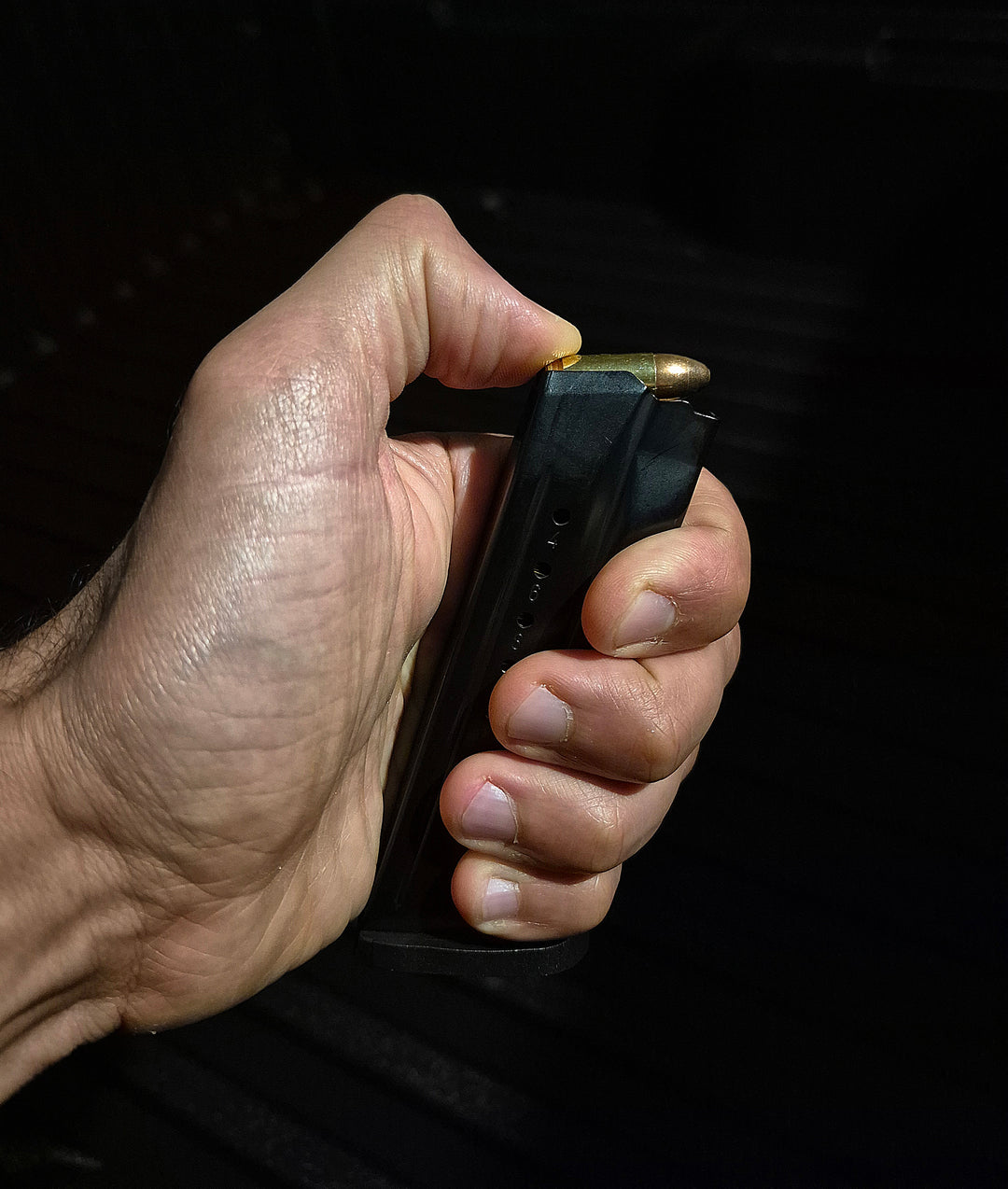Close-up of hand gripping loaded 9MM magazine against black background, showing brass-tipped rounds with textured grip and steel casing; styled for operational context and tactical realism to emphasize control, readiness, and component detail in live-use scenarios