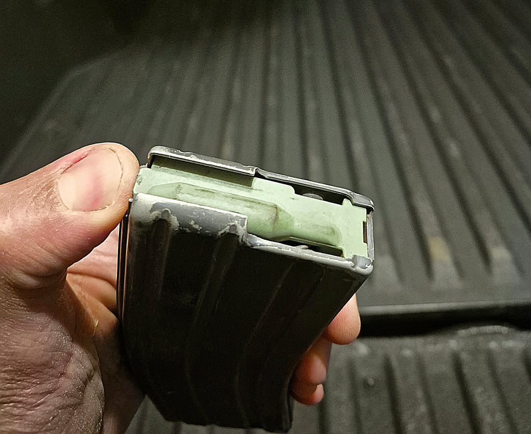 Hand holding DURAMAG aluminum magazine partially loaded with green dummy round, showing wear and field use