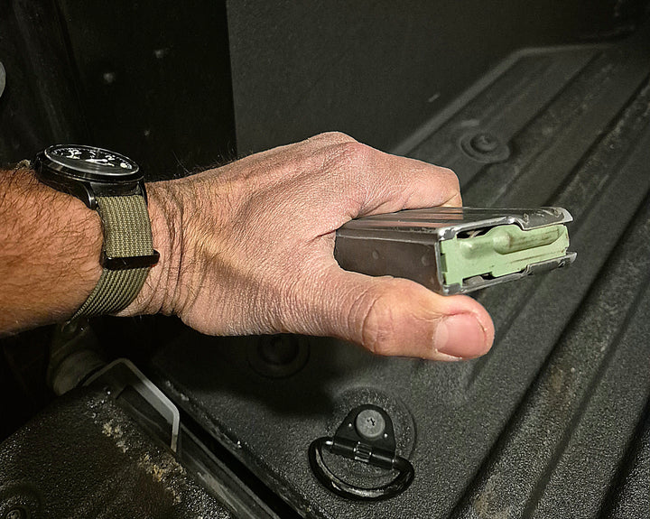 Hand holding DURAMAG aluminum magazine with green follower, shown against truck bed background
