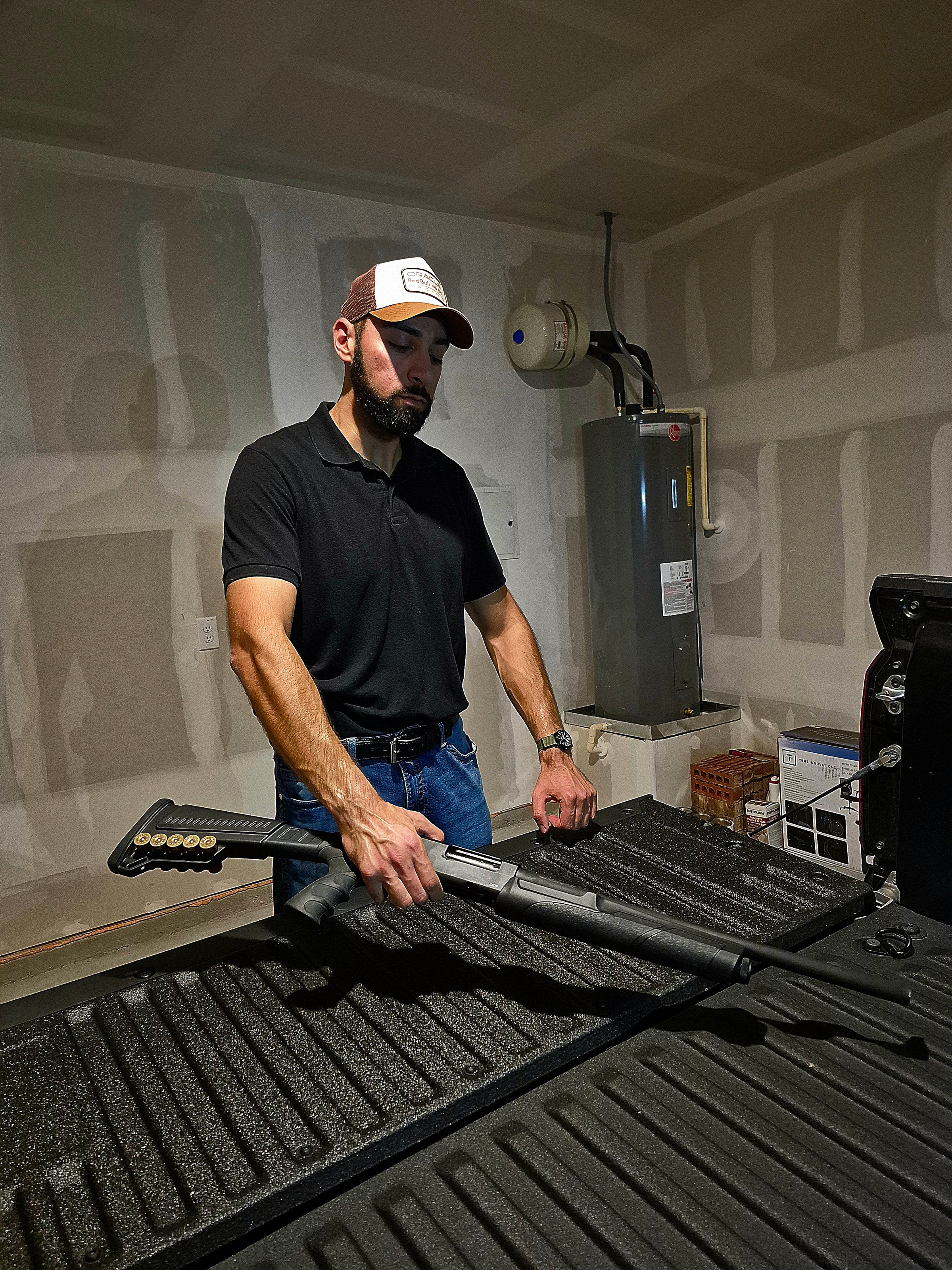 Sal Elboraa working on a vehicle bed cover inside a workshop, standing beside the truck bed while holding a tool; the space features exposed framing, a mounted water heater, and shelving with materials, suggesting a hands-on setup for tactical gear or product development