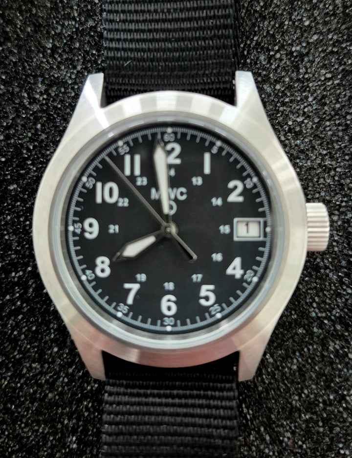 GS-002 MWC Classic Field Watch featuring a black dial with white numerals, silver bezel, and black NATO strap. Displayed on a textured surface in tactical lighting, highlighting its minimalist field-ready design.