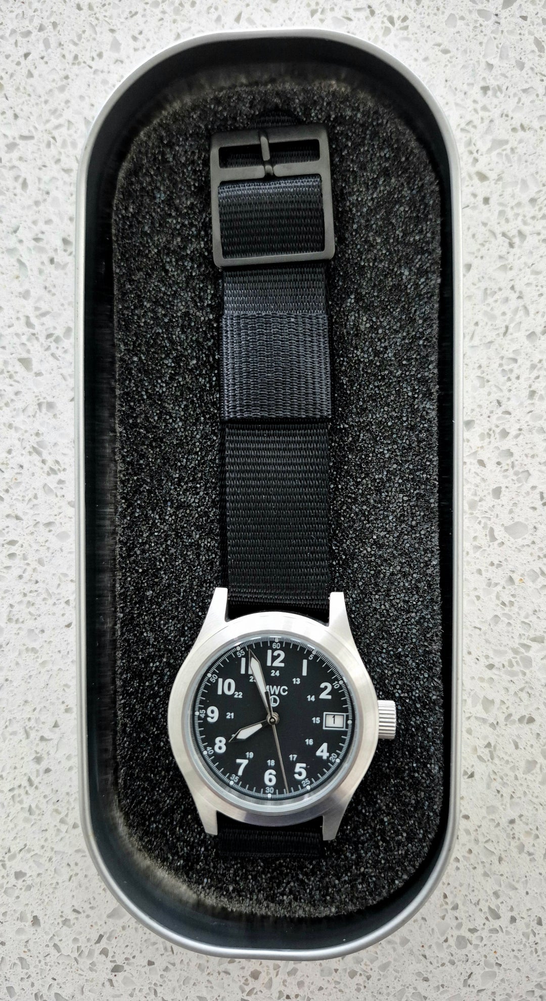 GS-002 MWC Field Watch with black dial and black strap inside a cushioned tactical watch box, displayed on a light gray surface for product staging.